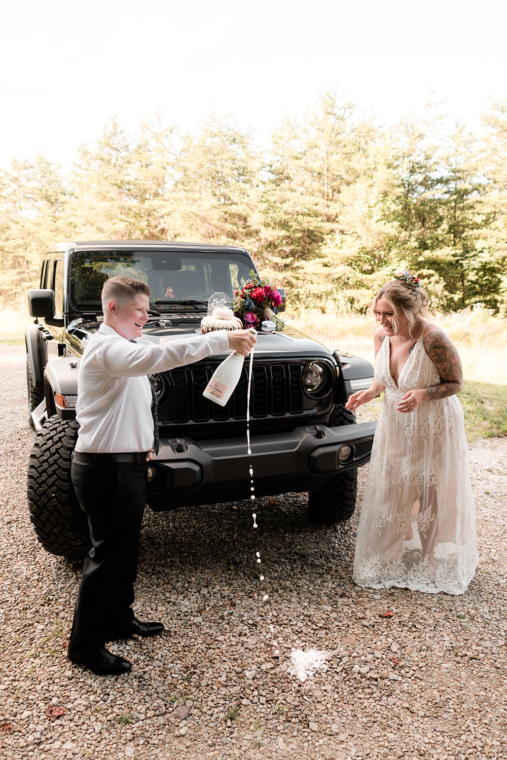 Two brides in wedding attire standing on a wooden cabin deck, laughing as they pop a bottle of champagne with forest trees in the background.