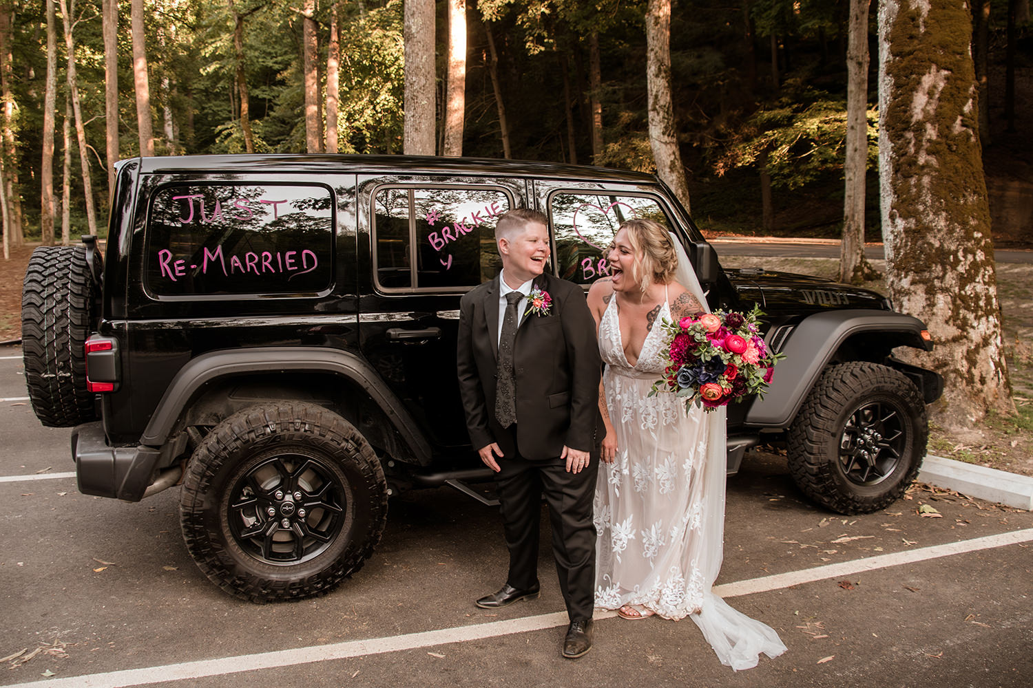 A queer couple laughs in front of their Jeep that has handwritten "Just Re-Married" text written on it with paint sticks.