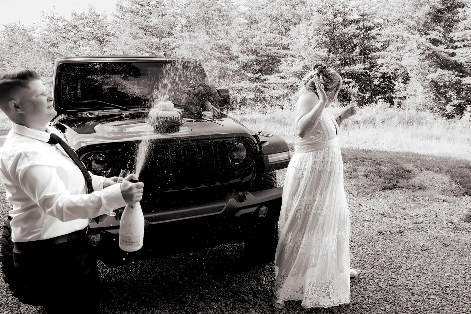 Two brides in wedding attire standing on a wooden cabin deck, laughing as they pop a bottle of champagne with forest trees in the background.