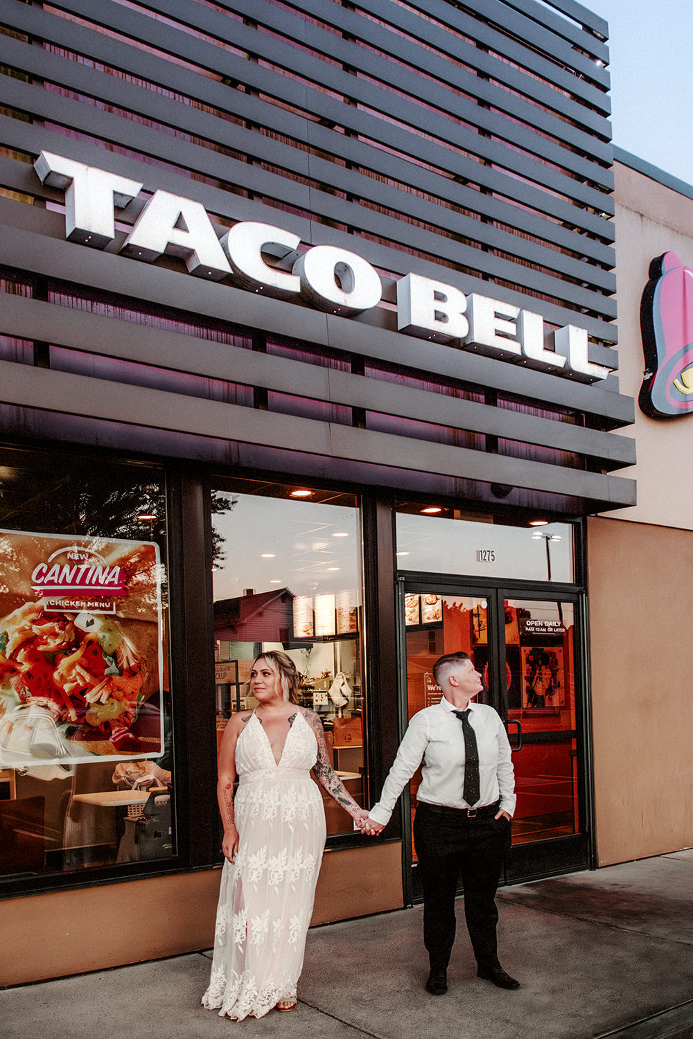Two brides hold hands and stand in front of a Taco Bell.
