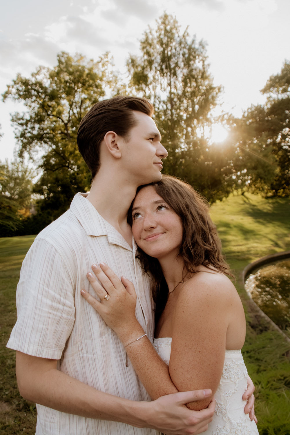 A woman presses herself against a man's chest and she looks up at him. He looks off into the distance. She wears a unique engagement ring. The sun is setting behind Schiller Park in the background.