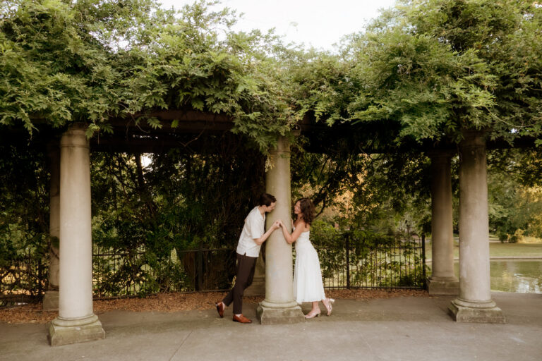 An engaged couple leaned inward on a column and they examine the woman's engagement ring. The column supports a cascade of green ivy. The location is Schiller Park in German Village, Ohio.