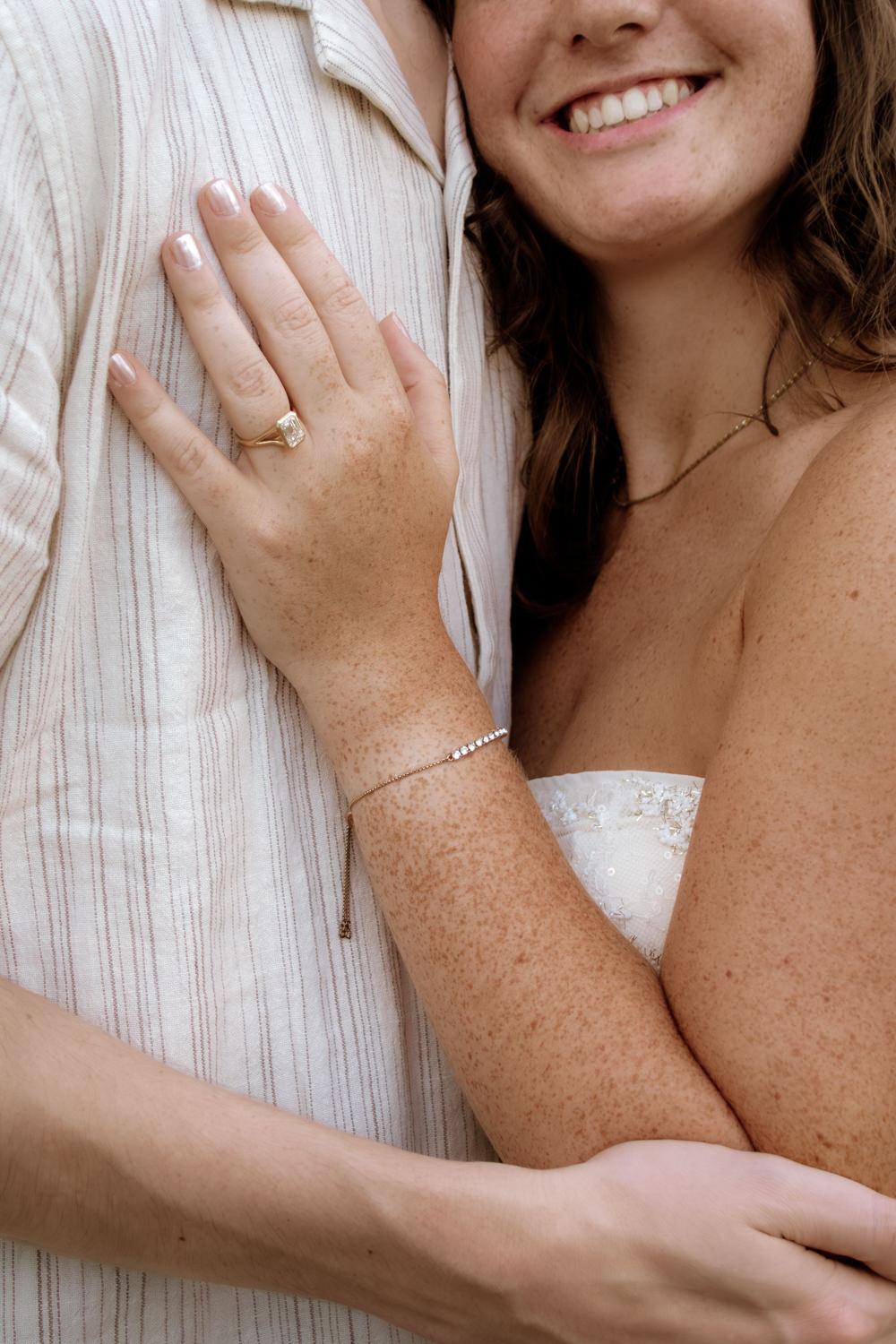 Close up crop of a woman's hand on a man's chest. She wears a unique engagement ring and diamonds bracelet. The photo is cropped just above her smile.