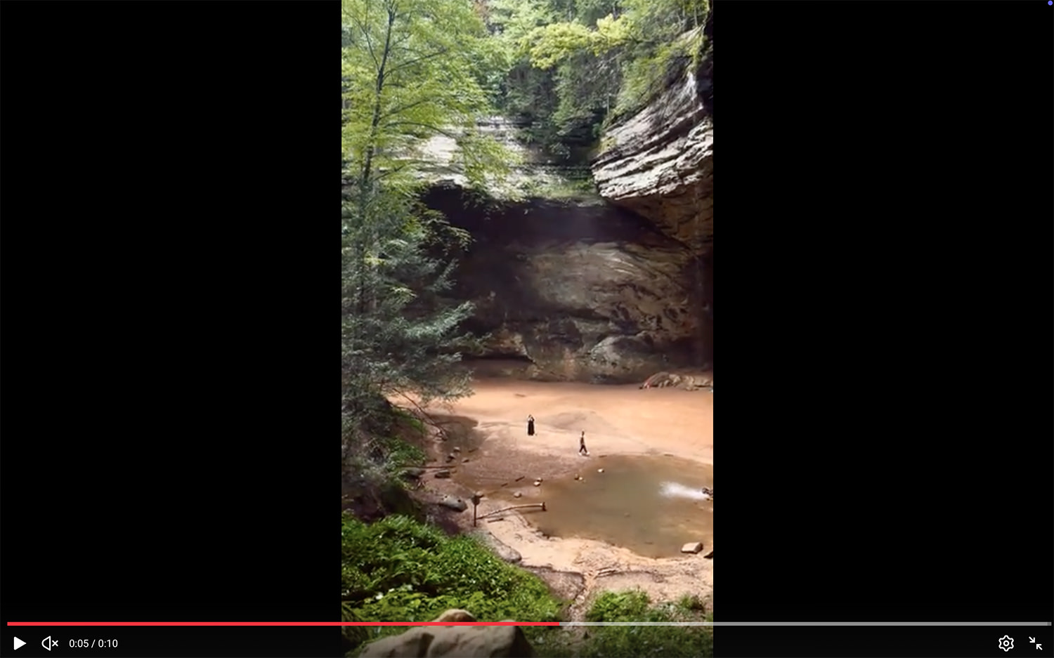 A screenshot of a video clip of two people by the waterfall pool at Ash Cave. The perspective is very wide and shows cliffs and the large cave.