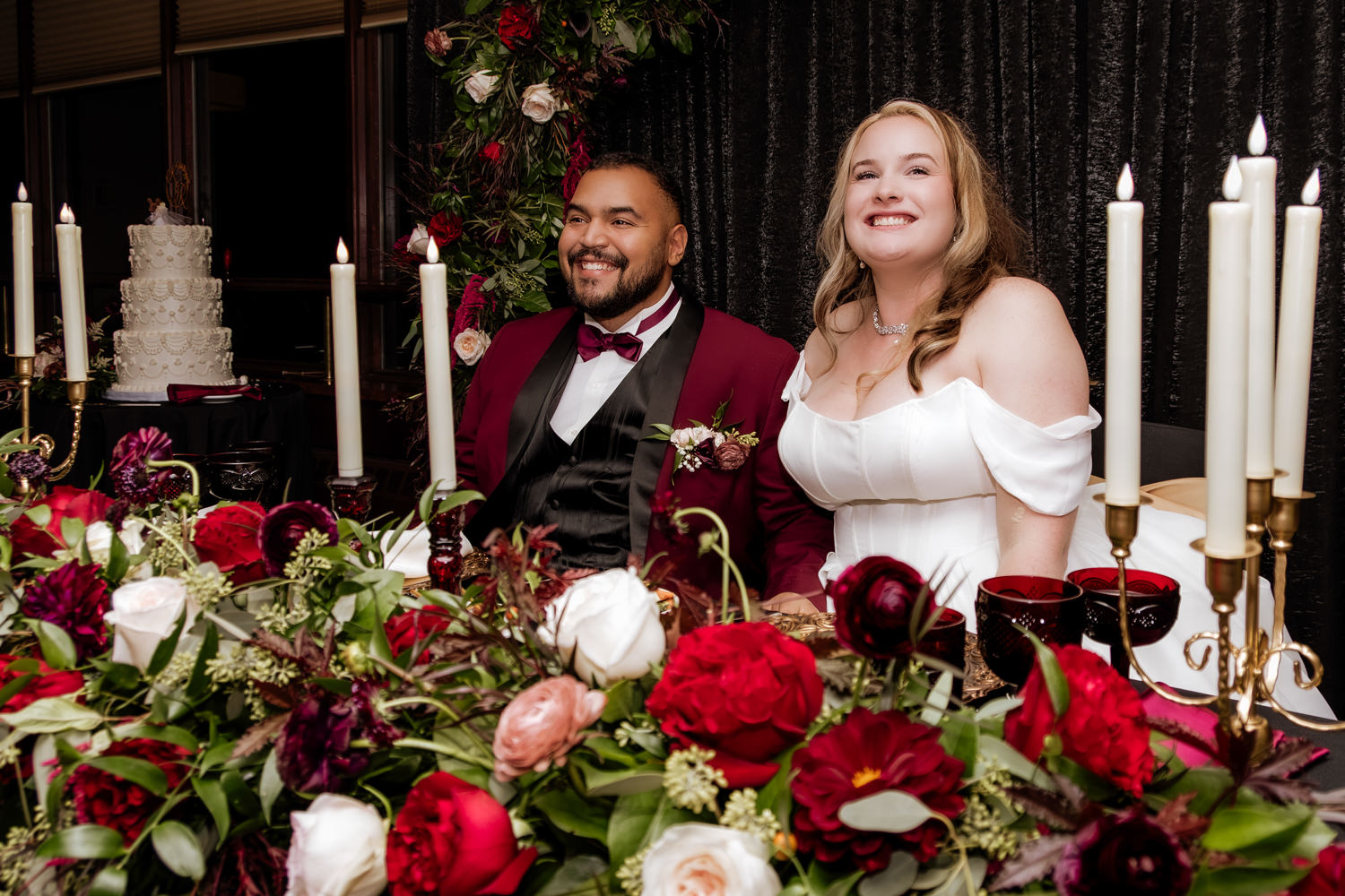 A newly married couple sitting at their intimate reception table with big smiles, surrounded by glowing candles and red autumn floral arrangements.