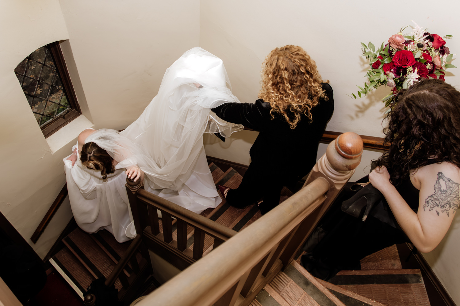 A high-angle perspective photo of a bride being carefully helped down a staircase by her mother and best friend before her autumn wedding.