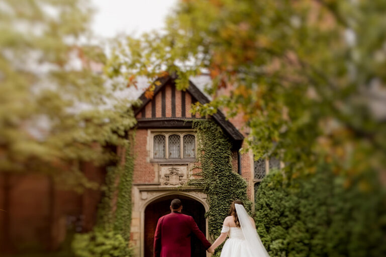 An artistically blurred photo of the backs of a couple walking towards a Tudor mansion entrance in fall. The groom is wearing a maroon suit. The bride has a flowing and dramatic veil.