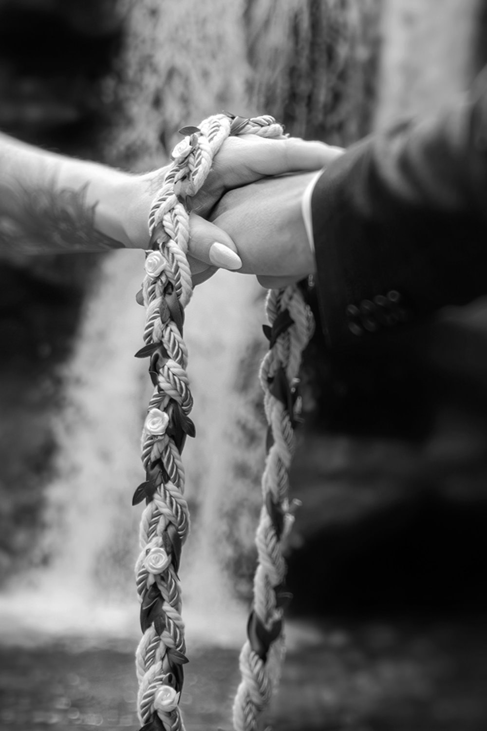 A close up of hands clasping and a braided hand fasting rope dangling in front of a waterfall. 
