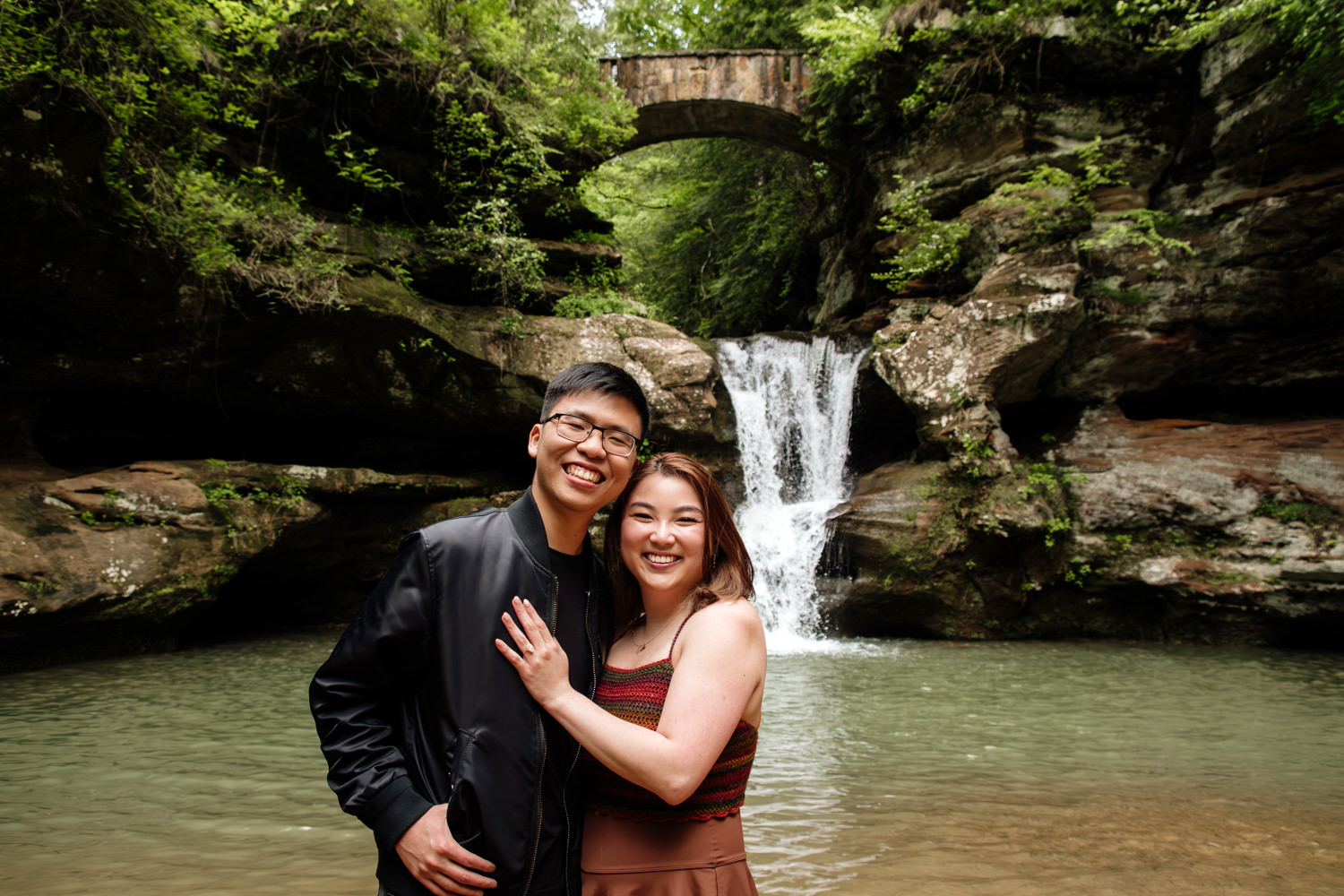 A smiling asian couple poses in front of Upper Falls after a proposal.