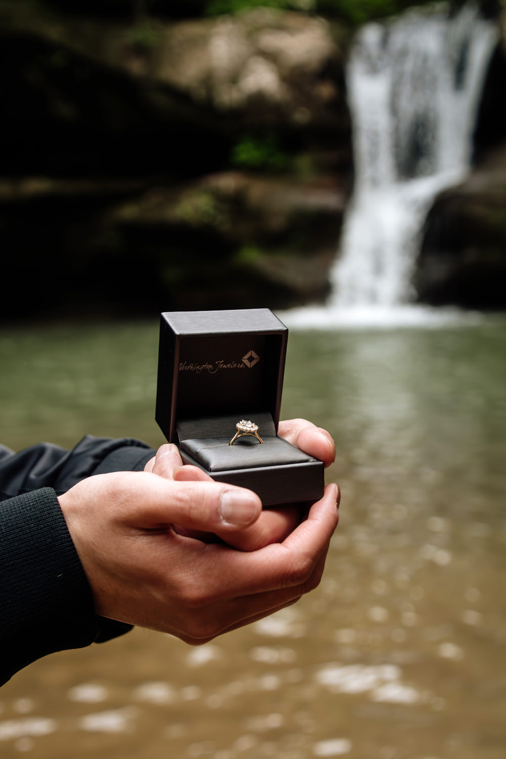 A man's hands clutch a ring box with an engagement ring.  In the background is a Hocking Hills waterfall and green pool of water.