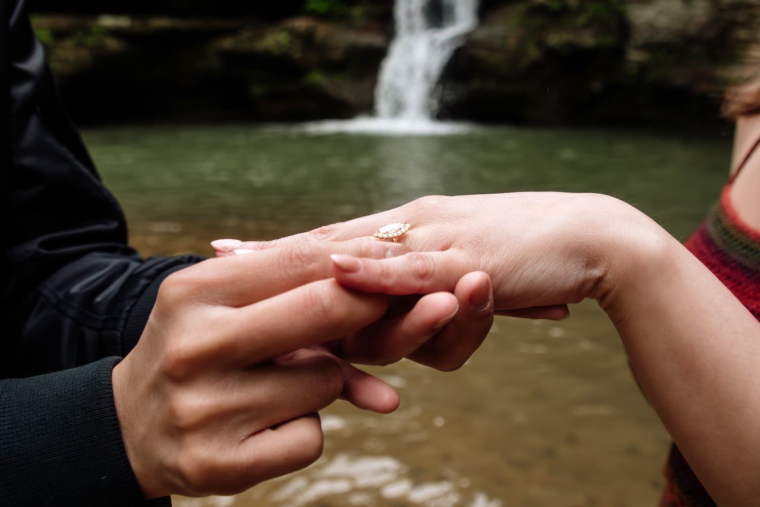 A man places an engagement ring on a woman's hand in front a of a Hocking Hills waterfall.