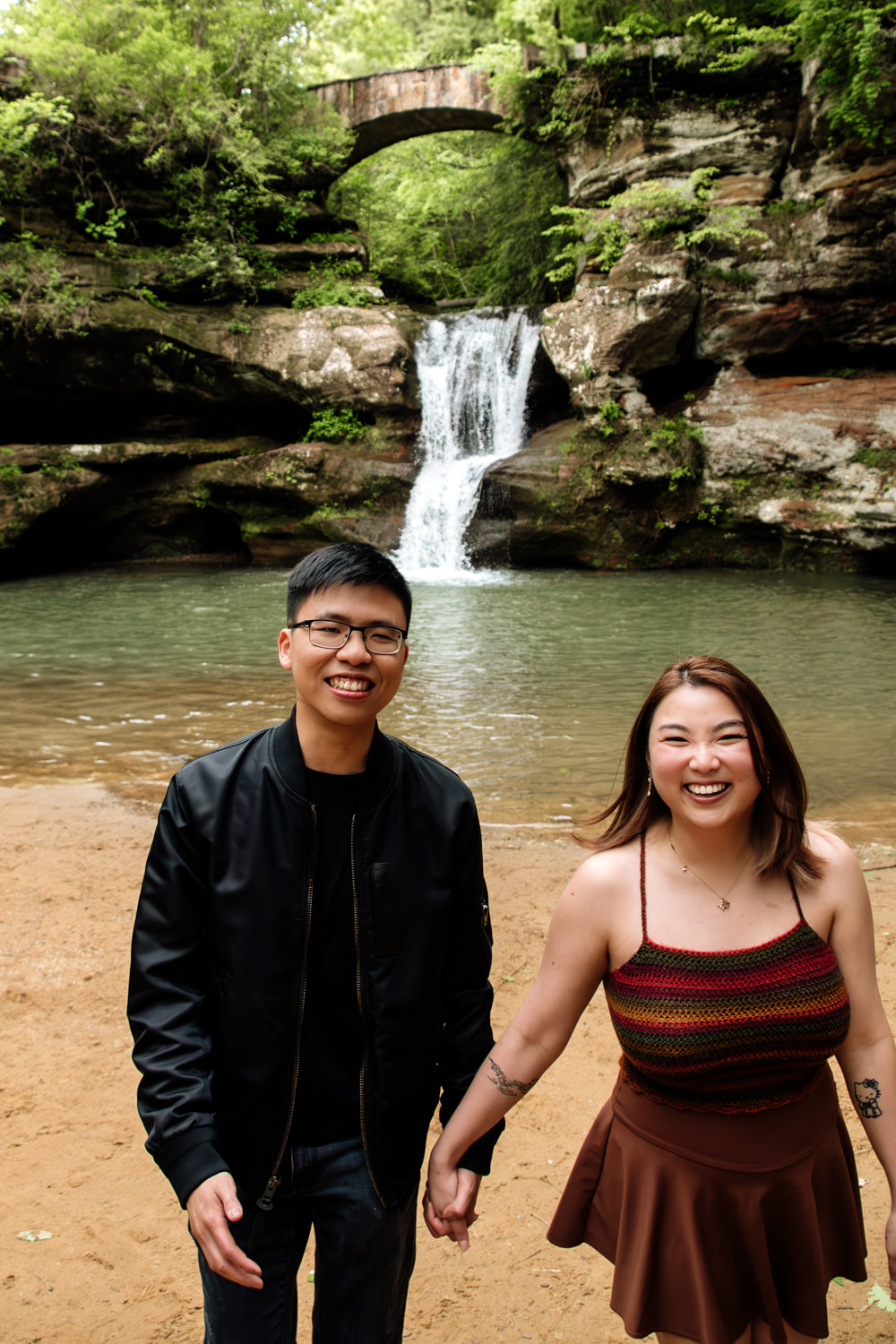 A couple holds hands and walks toward the camera and away from a waterfall in Hocking Hills.