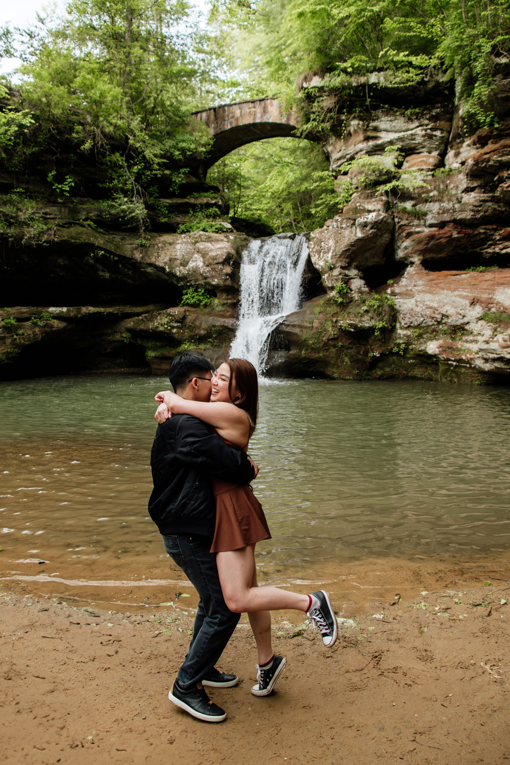An asian couple embraces and twirls in front of Upper Falls in the Hocking Hills after a surprise proposal.