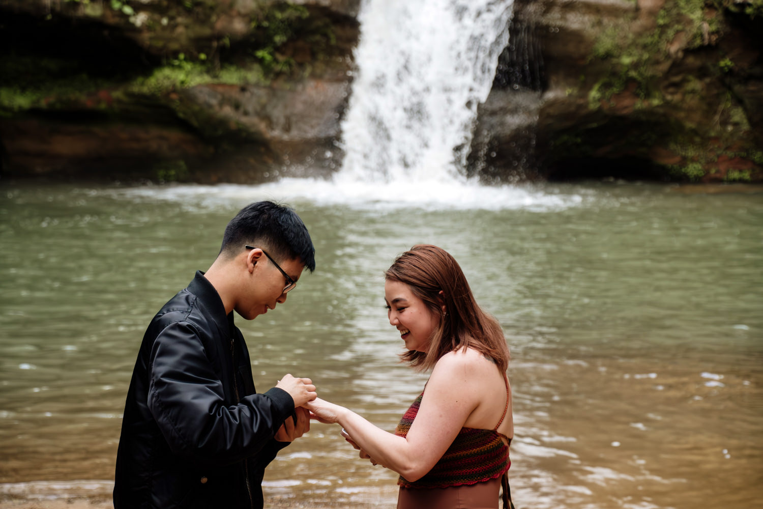 A man places an engagement ring on his girlfriend's finger. A waterfall rushes behind them.