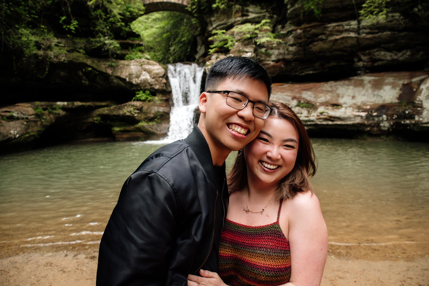 An asian couple smiles and poses in front of a Hocking Hills waterfall.
