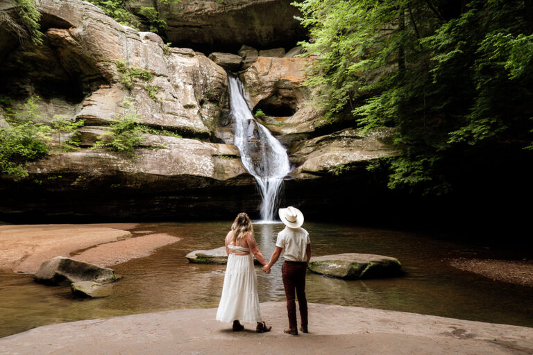 A couple standing on a Hocking Hills overlook of Cedar Falls. The woman wears a flowing cream-colored dress with cutouts, and the man wears an oatmeal-colored button-down with dark maroon pants. Their outfits complement each other with rich textures without being identical, standing out beautifully against the waterfall and sandstone backdrop.