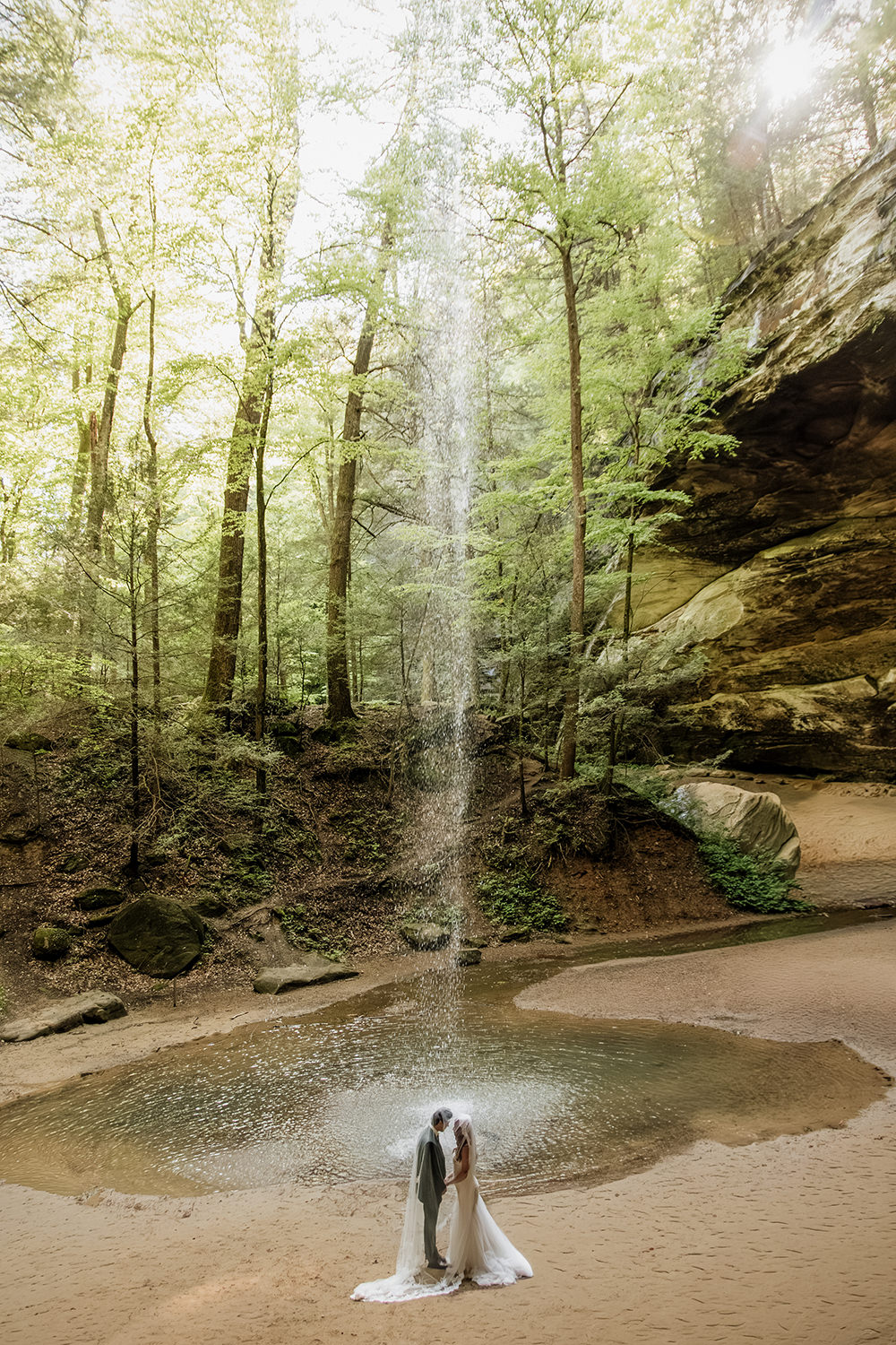 The Ash Cave waterfall flows from the overhang and drops down to where an elopement couple stands, small in scale, with a cathedral wedding veil draped over them.
