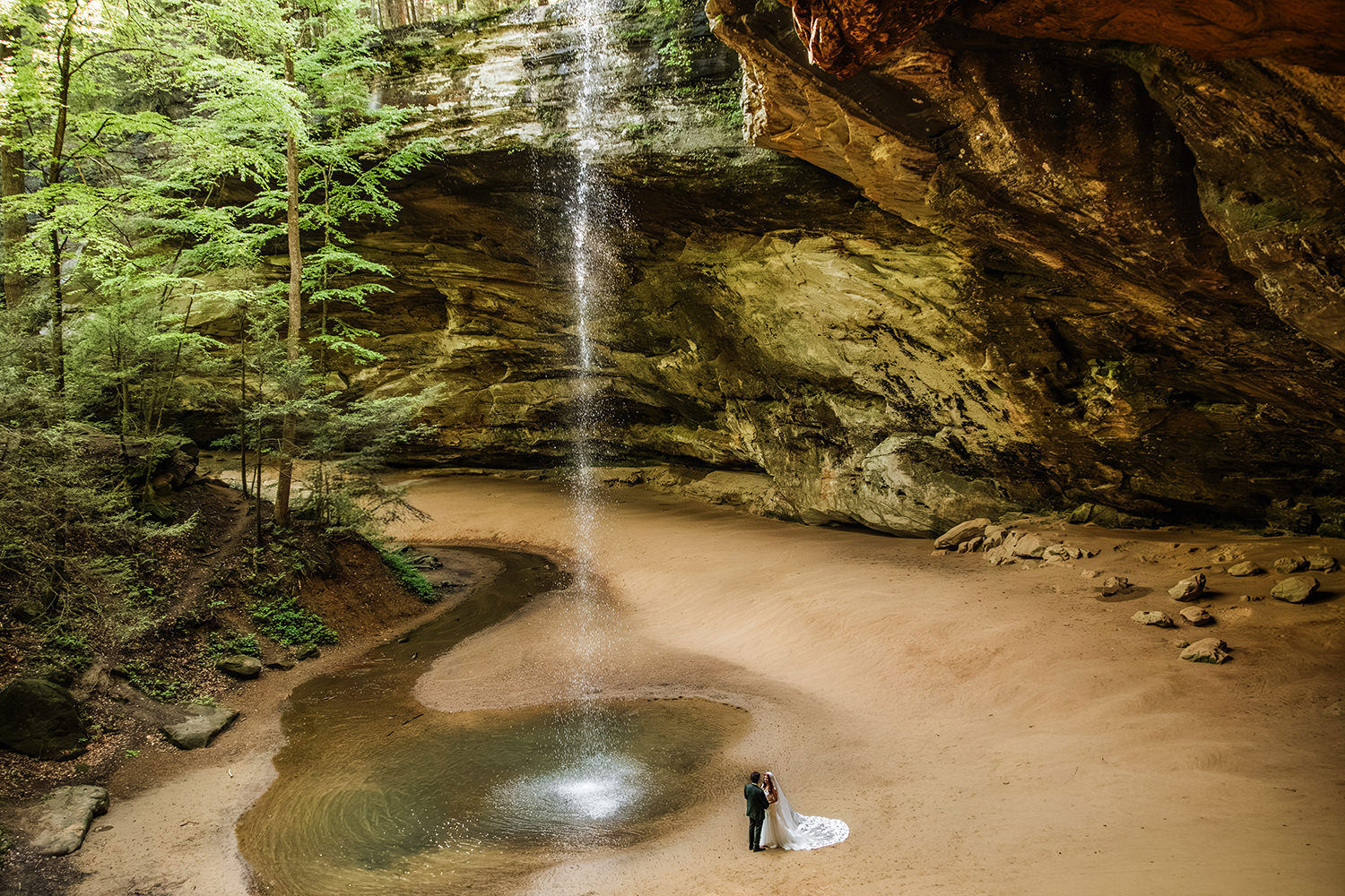 An epic, high-angle view looking down through a rushing waterfall at Ash Cave in Hocking Hills State Park, capturing an elopement ceremony with a bride and groom standing below.