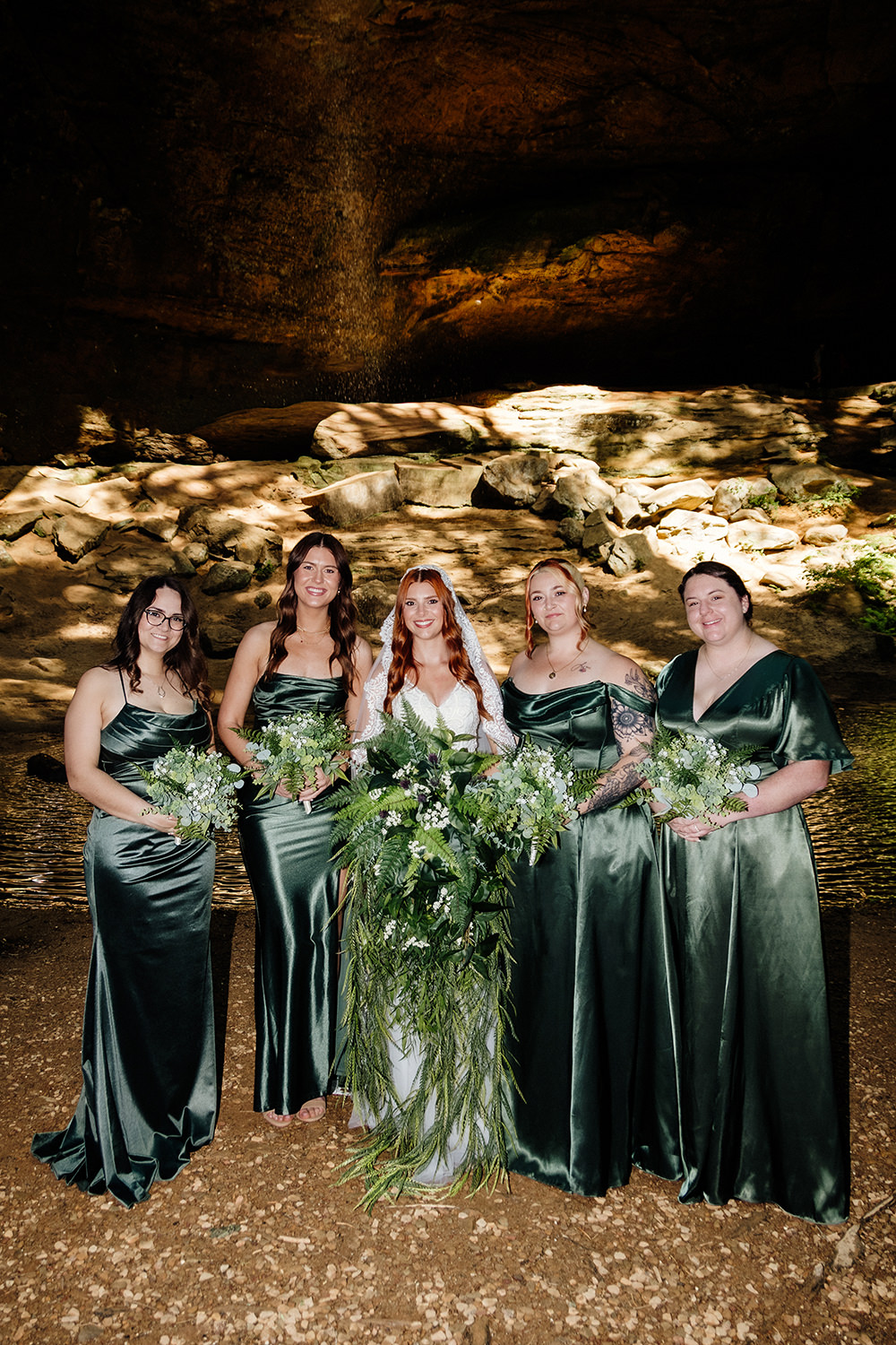 A group of bridesmaids in moss green dresses surround a bride wearing a vintage veil and red hair. The group is standing underneath a waterfall at Ash Cave.  The photo is captured with direct flash for a 90s nostalgic vibe.