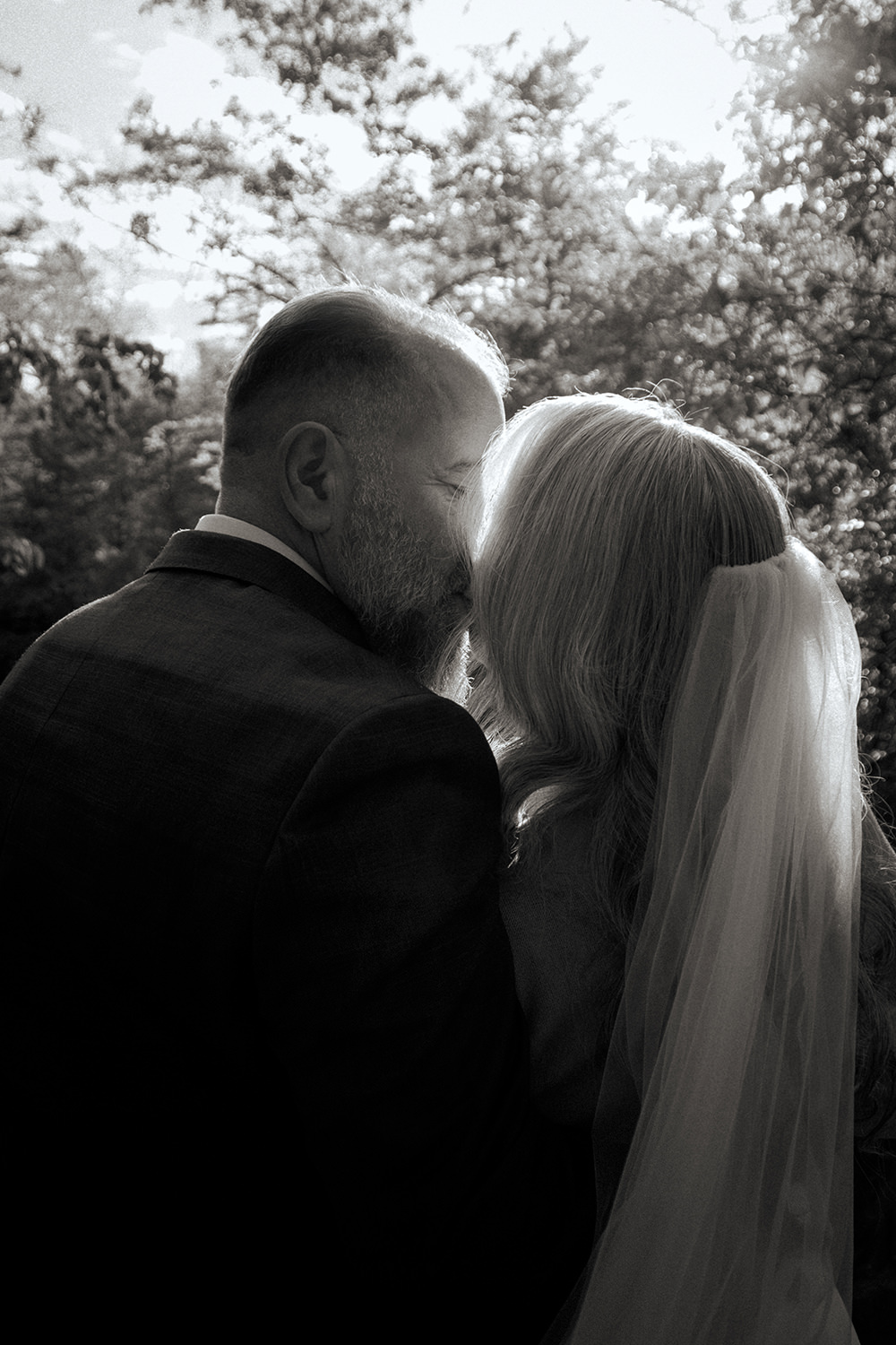 A black and white photo taken from behind an elopement couple as they kiss.  A glow gives them a ring light, illuminated their intimate moment.  