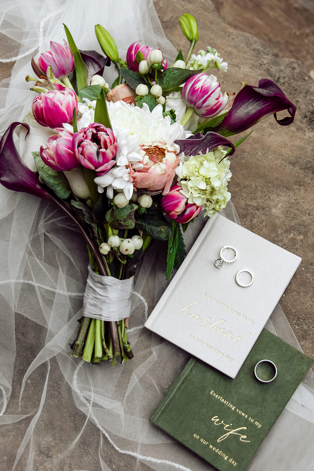 Flat lay photo of a wedding bouquet with white, pink, and purple.  Two vows books lie beside the bouquet with wedding rings on them.  