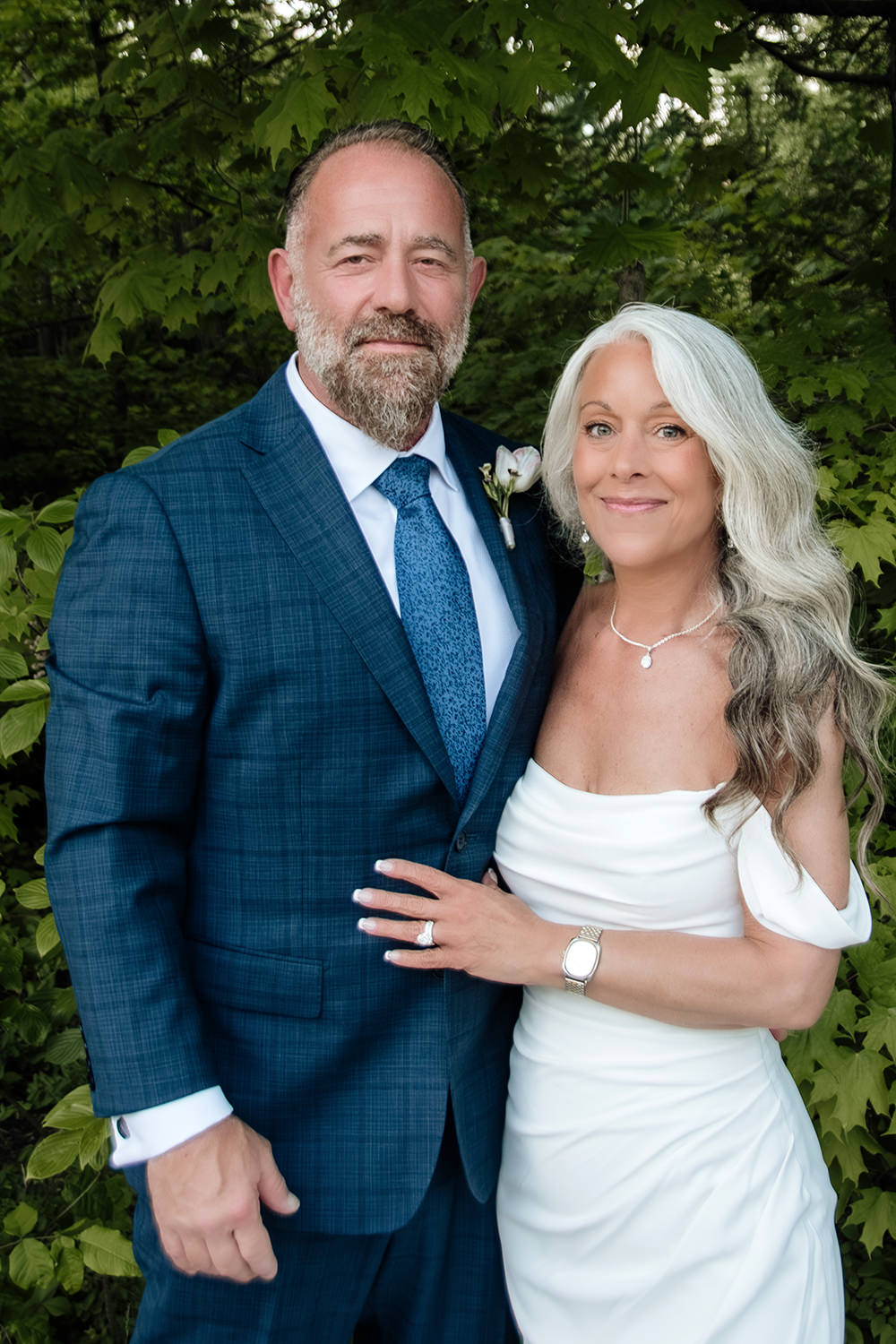 Traditional forward-facing portrait of an elopement couple looking at the camera and smiling.  The man wears a blue suit, has a salt and pepper beard, and is in his 60s. The woman has long white hair and wears an off-the-shoulder mermaid wedding gown.