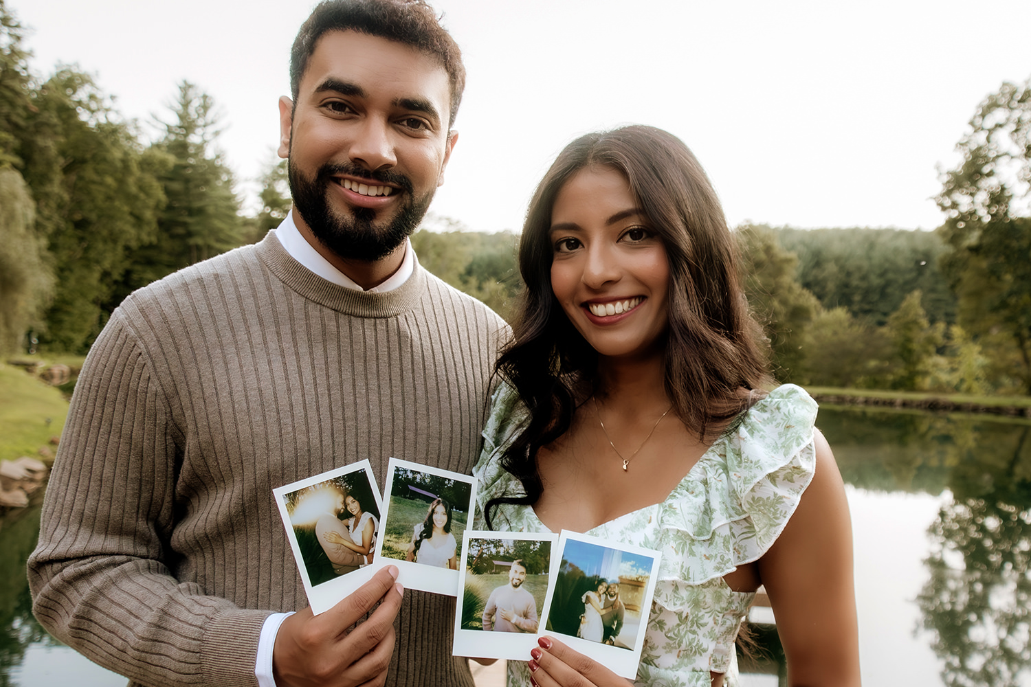 A couple wearing neutral browns, sage green, and cream smile at the camera and hold four Polaroids.  In the background, the sun sets over a pond and hills.