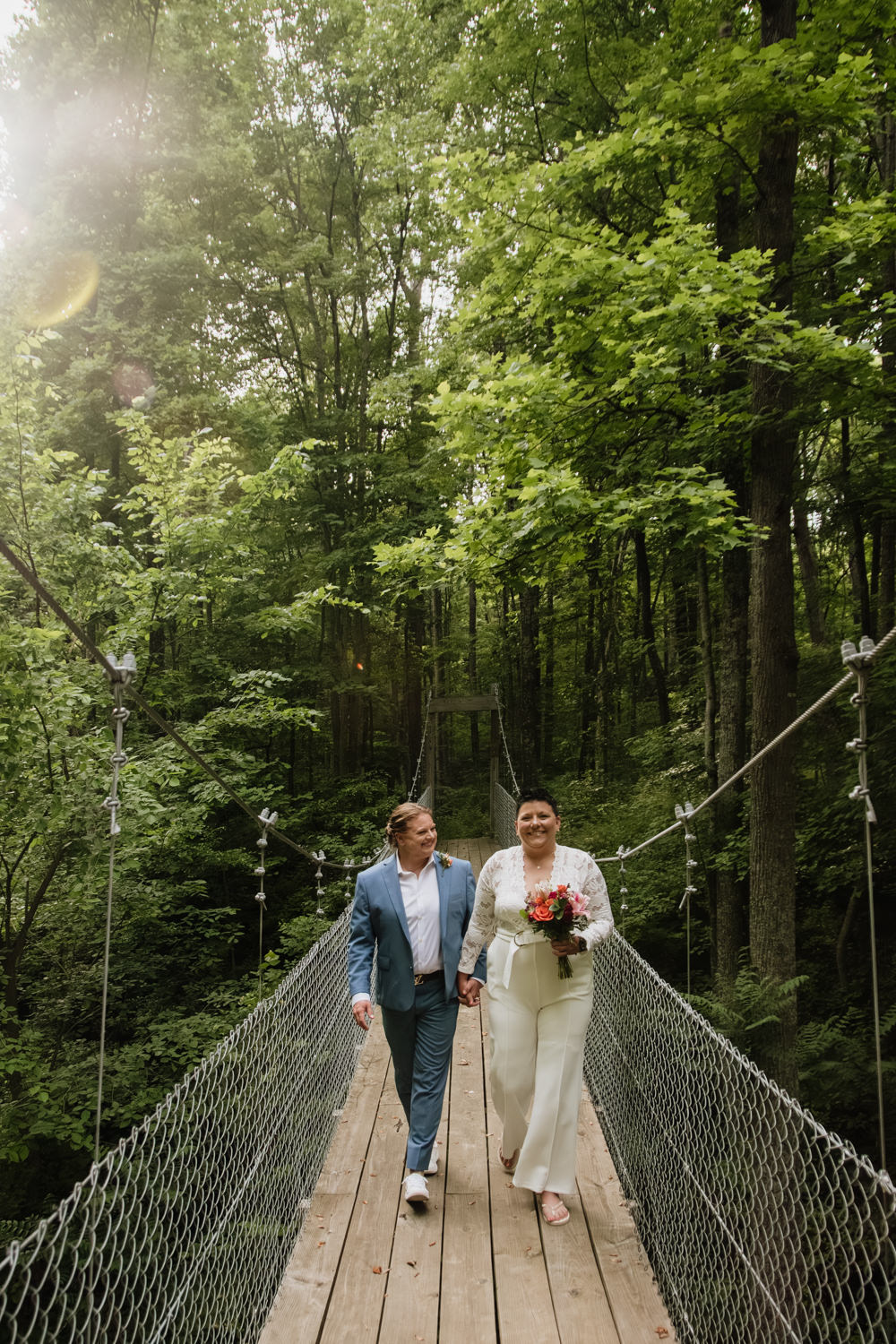 Wide view of a couple standing in the middle of a wooden suspension bridge surrounded by deep green forest.