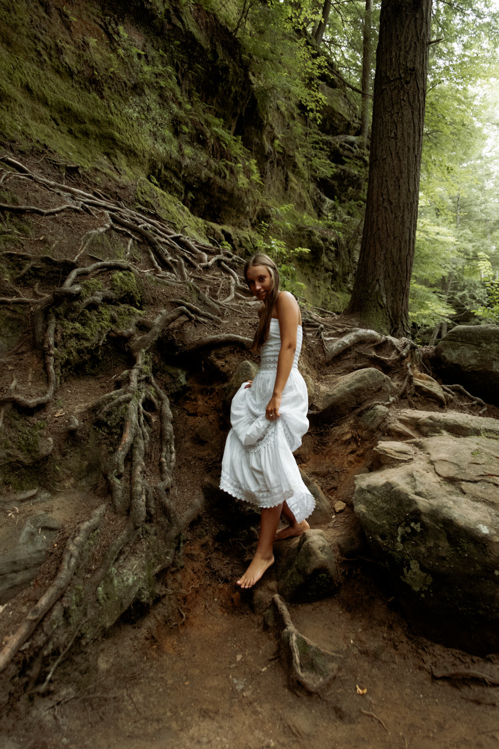 High school senior in a flowing cream dress walking along a sandstone trail in Hocking Hills.