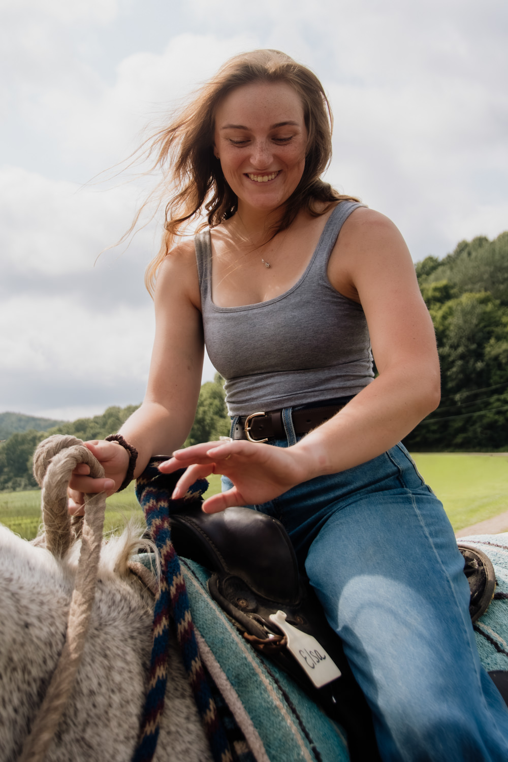 A woman sits in a saddle on a trail ride and examines her new engagement ring while she smiles big.