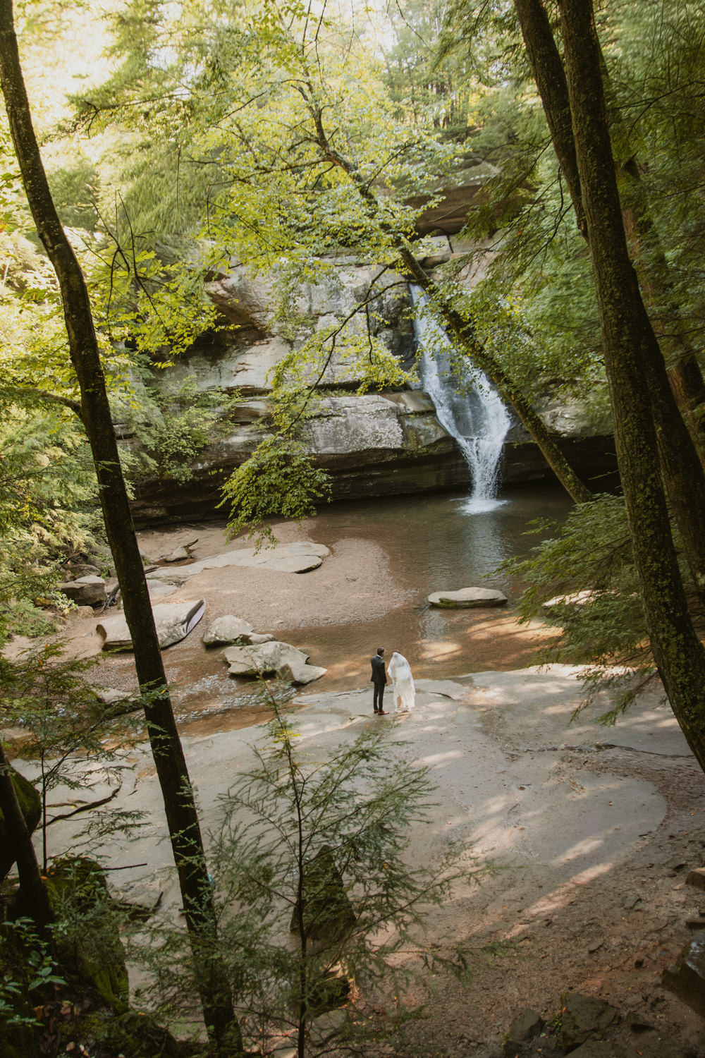 Bride and groom holding hands in front of a full-flowing waterfall in August.