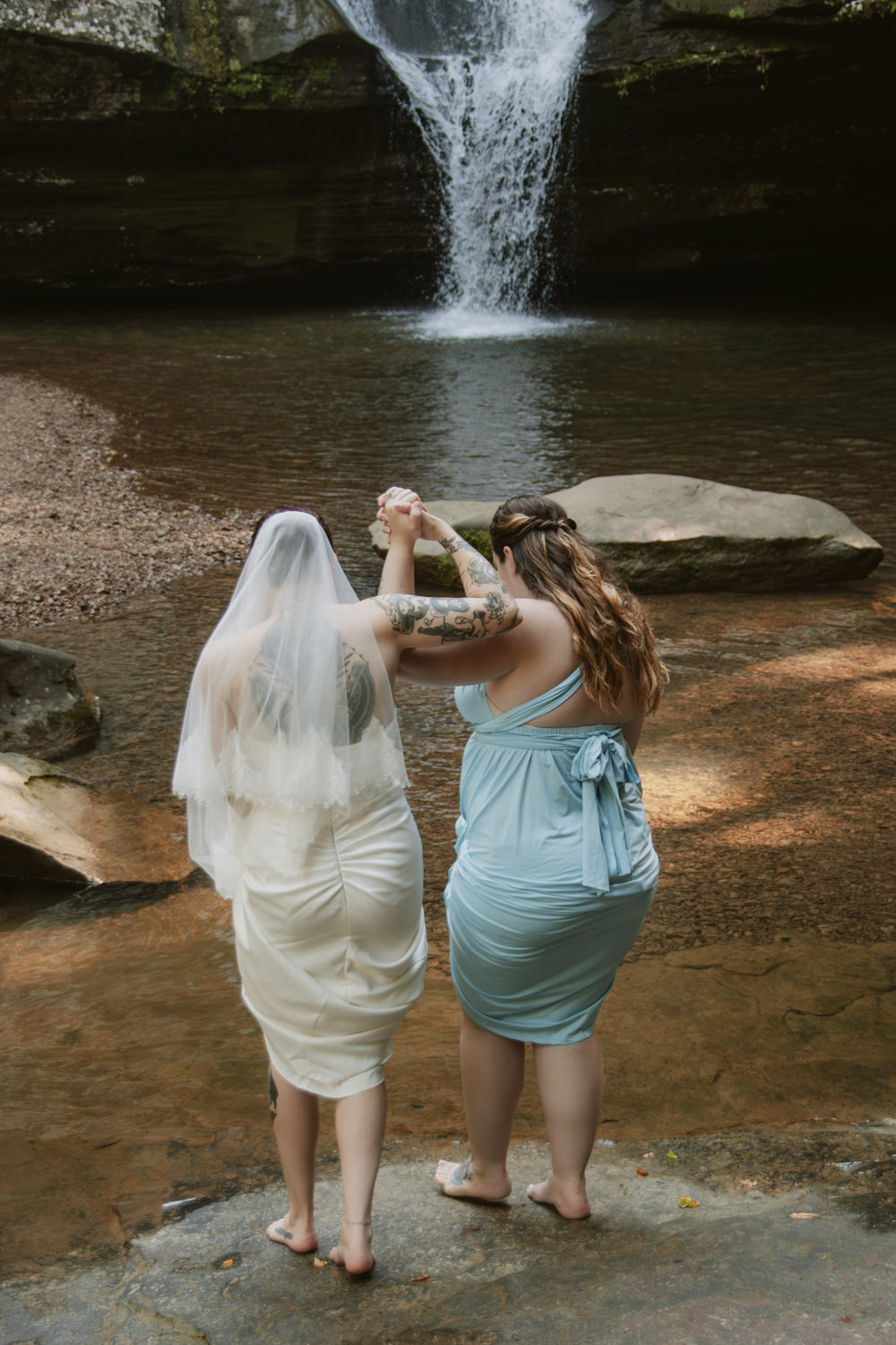 A maid of honor helps a bride into a creek in front of Cedar Falls.