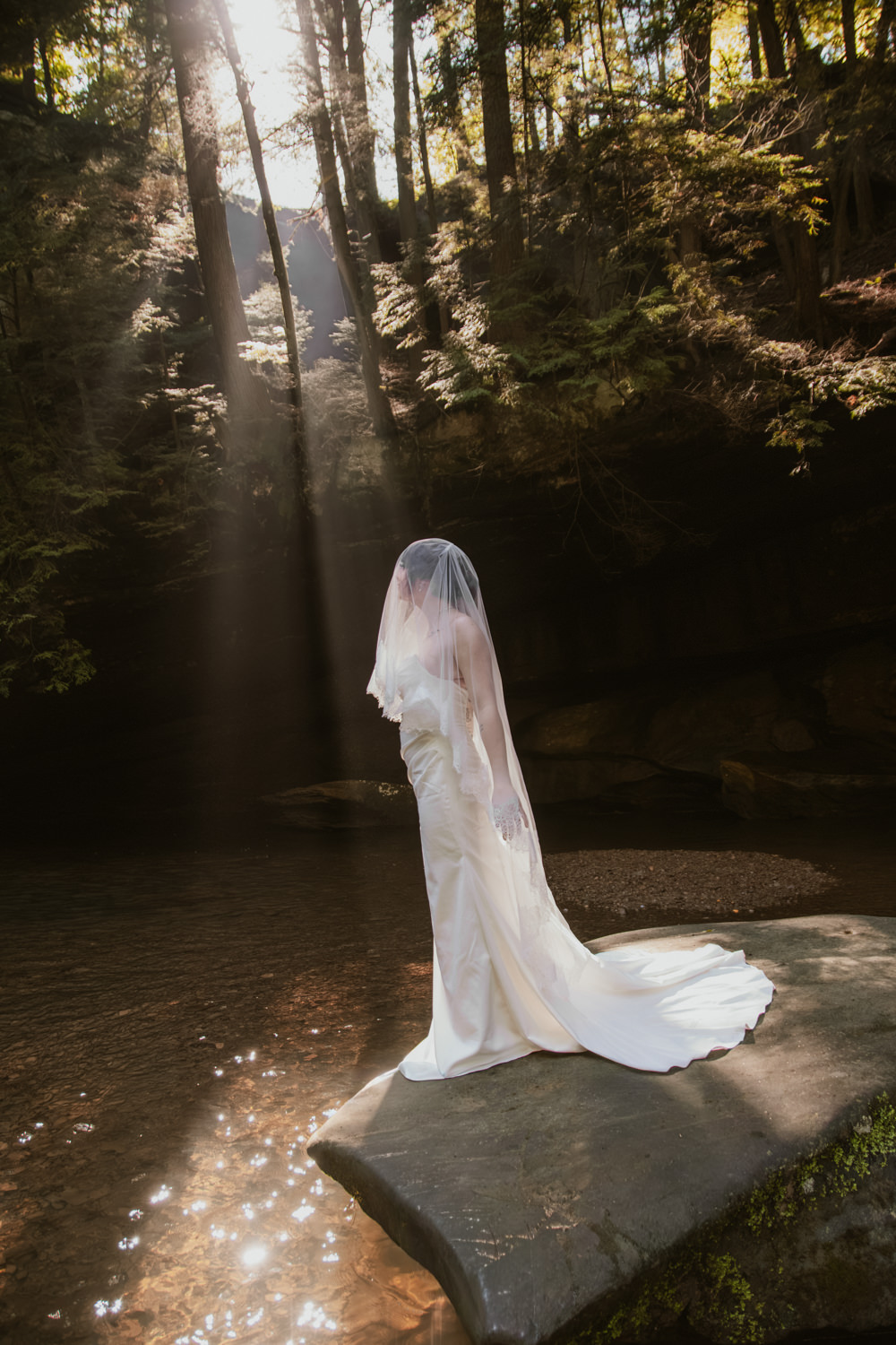 Bride in her white dress standing atop a giant mossy boulder illuminated by a beam of sunlight.