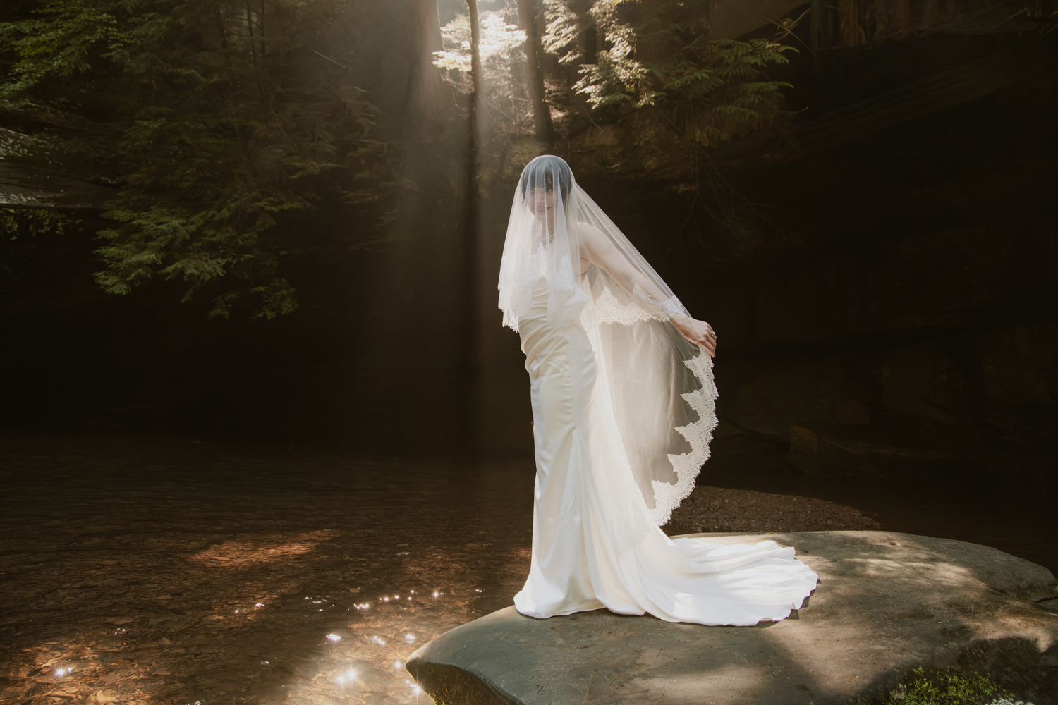 Bride in her white dress standing atop a giant mossy boulder illuminated by a beam of sunlight.