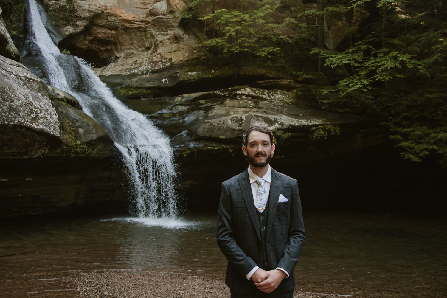 A groom poses in front of a flowing waterfall in Hocking Hills.