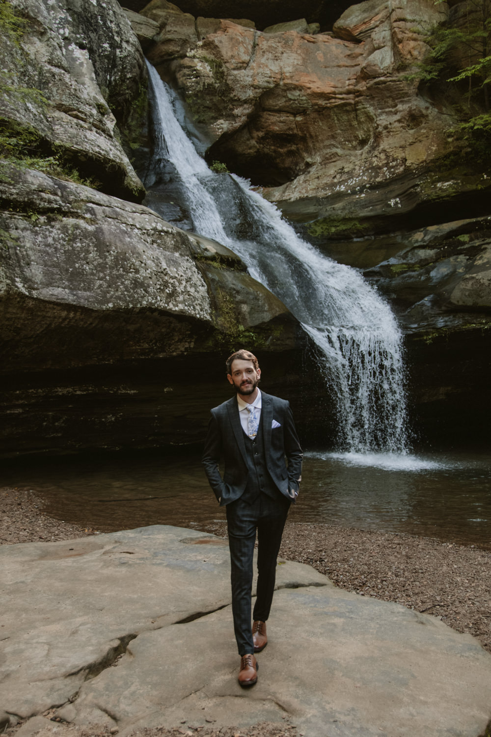 A groom poses in front of a flowing waterfall in Hocking Hills.