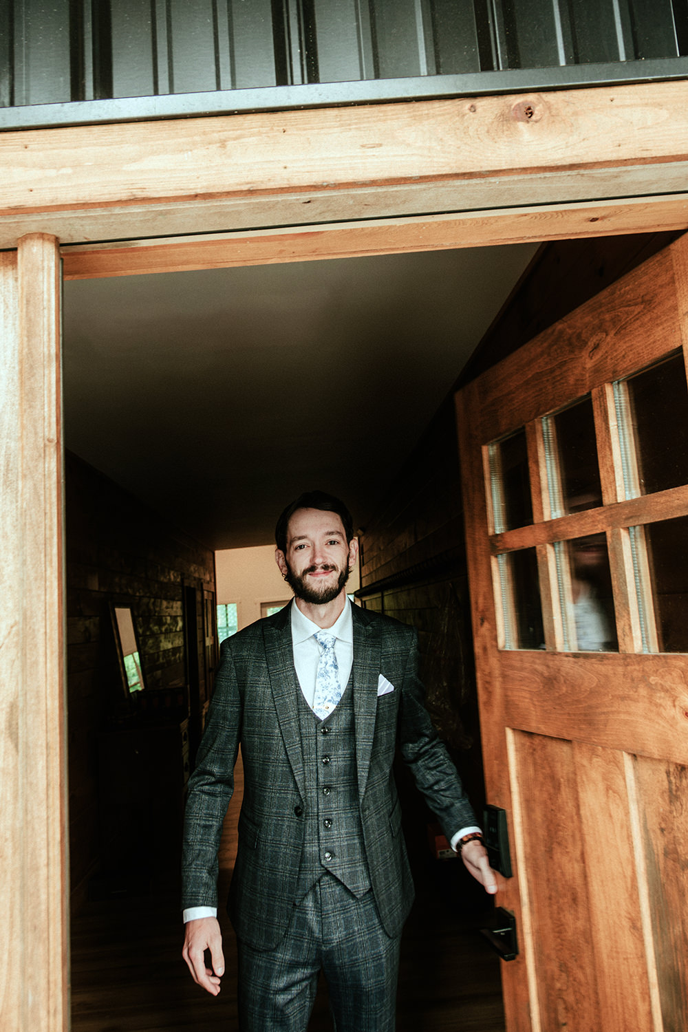 Groom in a dark suit tying his tie in front of a warm cabin window.