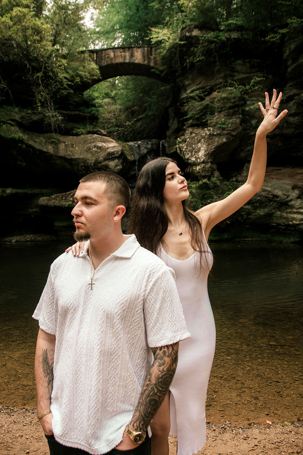 Soft golden sunlight filtering through forest leaves onto the couple standing near the waterfall area.