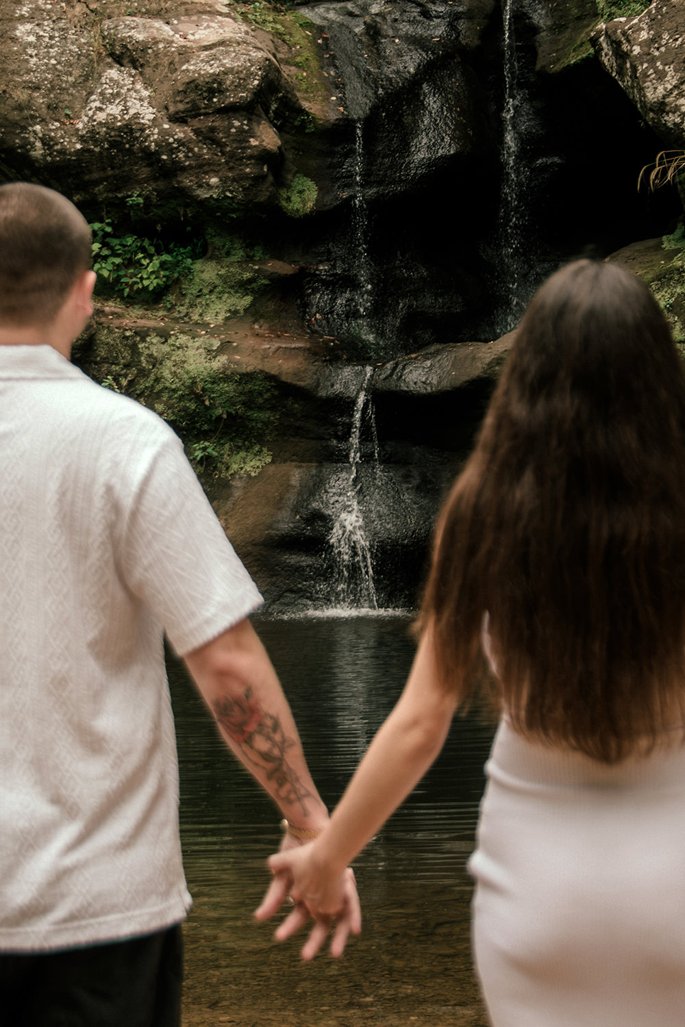 An engaged couple stands out of focus and holding hands in front of Upper Falls at Old Man's Cave.