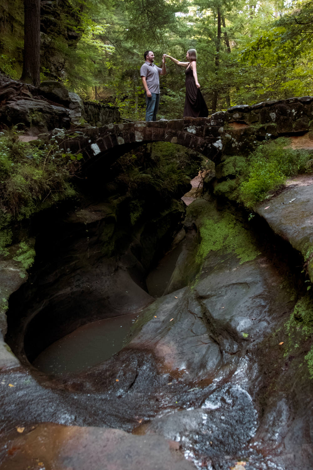 Couple standing on a stone bridge surrounded by lush green forest in Hocking Hills, Ohio. The bridge overlooks the Devil's Bathtub.