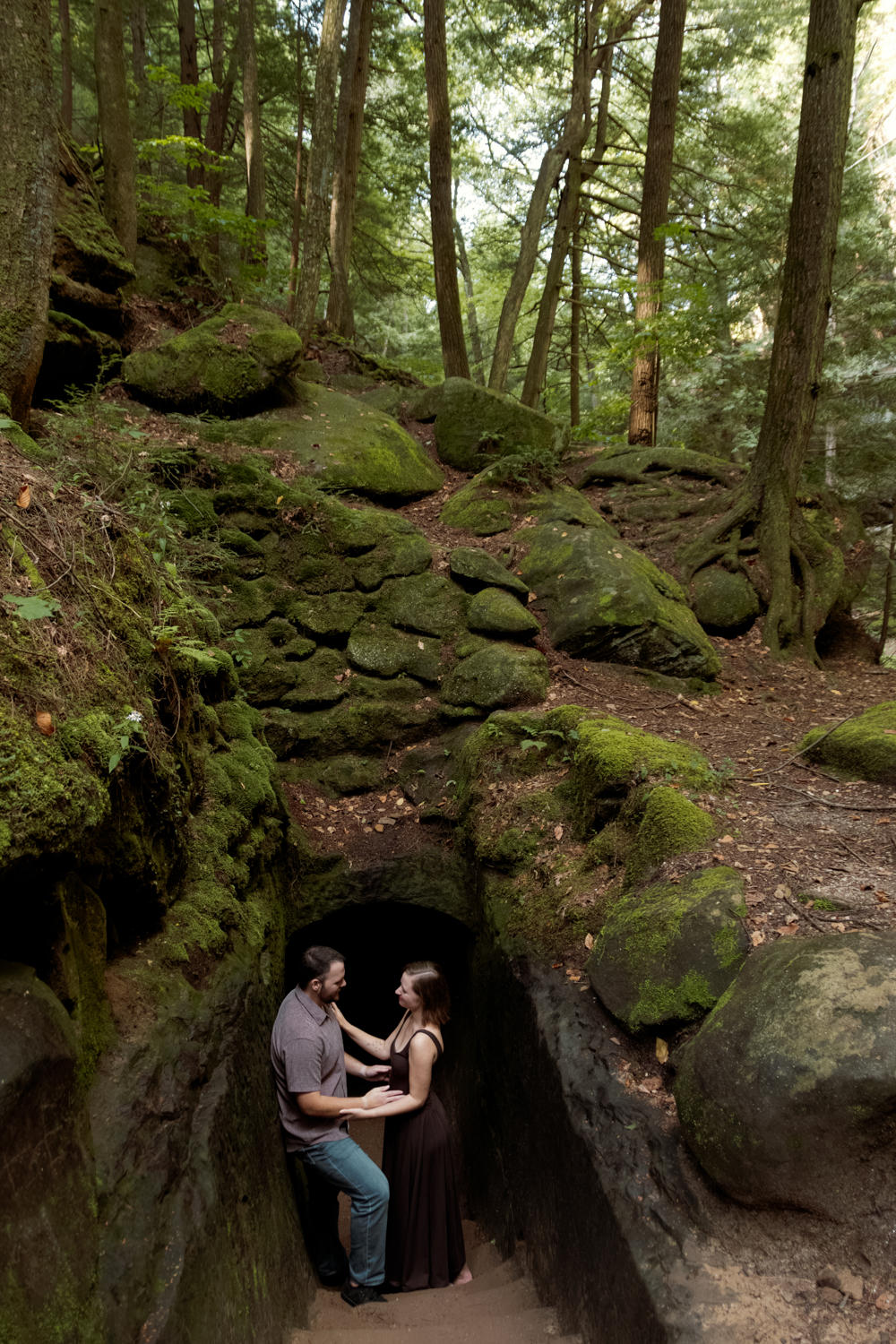 Sunlight breaking through the thick tree canopy onto a happy couple standing at the entrance of a tunnel in the woods.