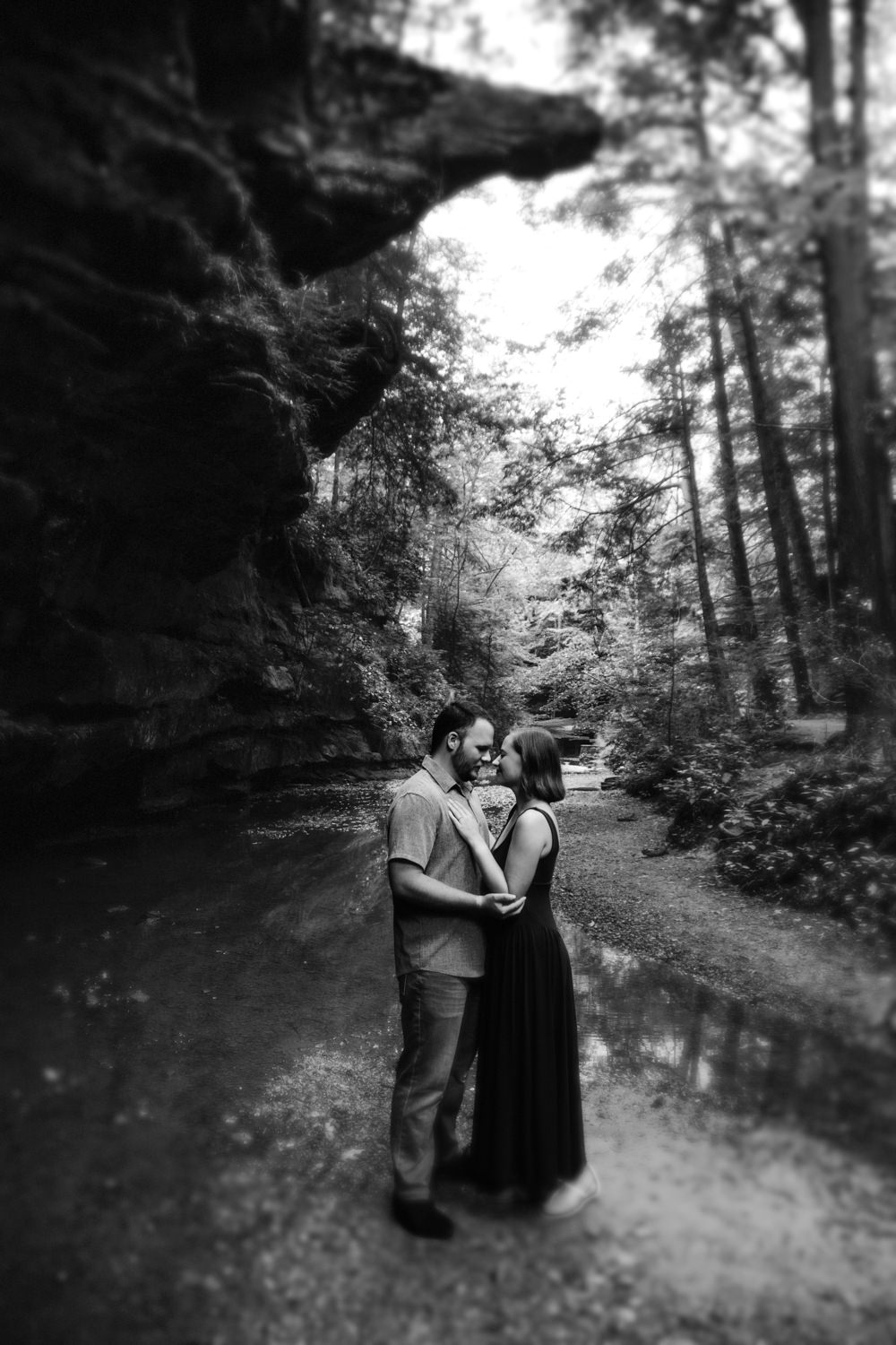 Black and white portrait of a couple kissing beneath a giant sandstone rock overhang in the forest.